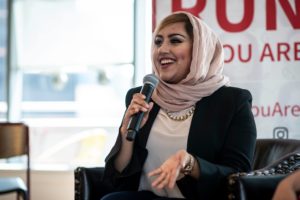 A woman wearing a light pink hijab laughs while holding up a microphone to her mouth.