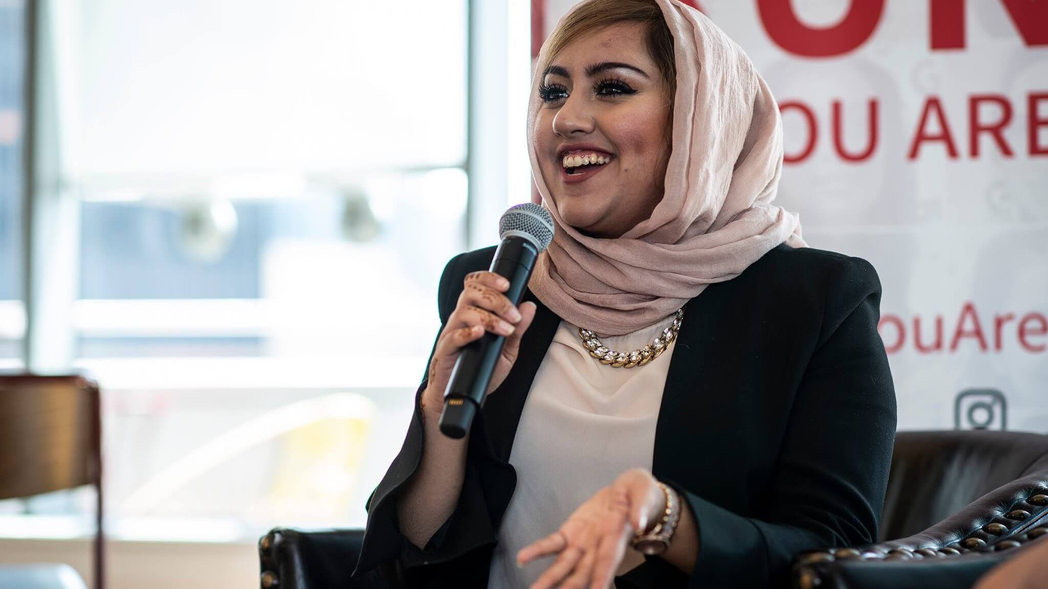 A woman wearing a light pink hijab laughs while holding up a microphone to her mouth.
