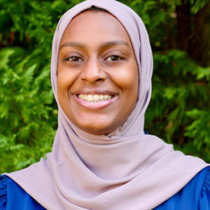 A young Black woman wearing a mauve headscarf and blue top smiles in front of green leaves.
