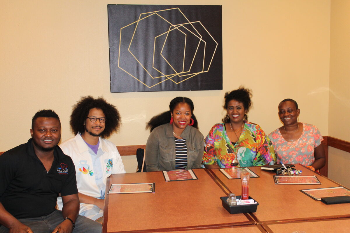 Lovely Grille chef sitting and smiling facing the camera with WFM staff, Fowzia and Vivian